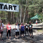 Osiem osób z kijkami do nordic walking czeka pod banerem z napisem start. Po prawej stronie sędzia daje pistoletem sygnał startu.