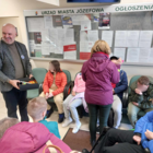 Gruba osób niepełnoletnich w okryciach wierzchnich, siedzących w holu Urzędu Miasta oraz stojący mężczyzna ubrany w ciemny golf i marynarkę, trzymający w rękach pudełko z odblaskowymi zawieszkami.