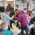 Gruba osób niepełnoletnich w okryciach wierzchnich, siedzących w holu Urzędu Miasta oraz stojący mężczyzna ubrany w ciemny golf i marynarkę, trzymający w rękach pudełko z odblaskowymi zawieszkami.