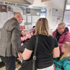 Gruba osób w okryciach wierzchnich w tym młody chłopak na wózku inwalidzkim, oczekujących w holu Urzędu Miasta oraz stojący tyłem mężczyzna ubrany w ciemną marynarkę.